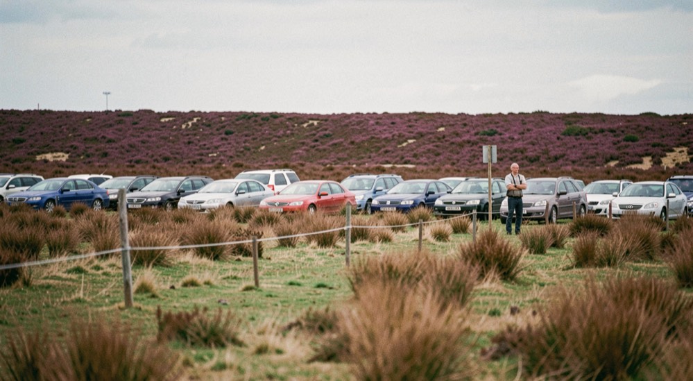 A wide moorland trial field in late summer with heather on the skyline, a line of parked cars along a rope and a shepherd alone at the post